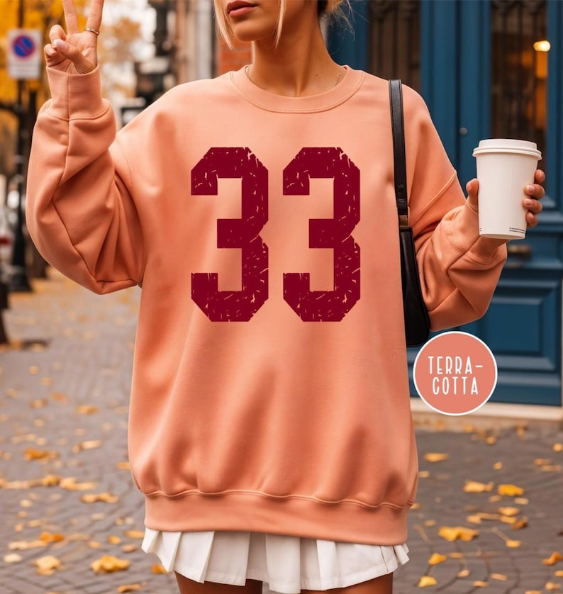 Op de afbeelding: Een perzikkleurige sweatshirt met het nummer 33 in een verweerde kastanjebruine lettertype. De sweatshirt heeft een ronde hals en lange mouwen. Een witte geplooide rok is zichtbaar. Een klein rond logo met de tekst "TERRA-COTTA" is zichtbaar.