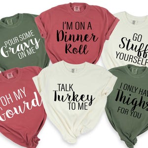 May include: Six short-sleeved t-shirts in shades of green, red, and cream, each printed with a Thanksgiving-themed slogan in a contrasting font. Phrases include "Go stuff yourself" and "Talk turkey to me."