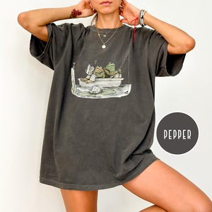 Op de afbeelding: Donkergrijze oversized t-shirt met een grillige illustratie van twee kikkers in een boot, een lezend en de ander aan het vissen. De shirt heeft een relaxte pasvorm en een vintage wassing. Het woord "PEPPER" is gedrukt op een donkergrijze cirkel.