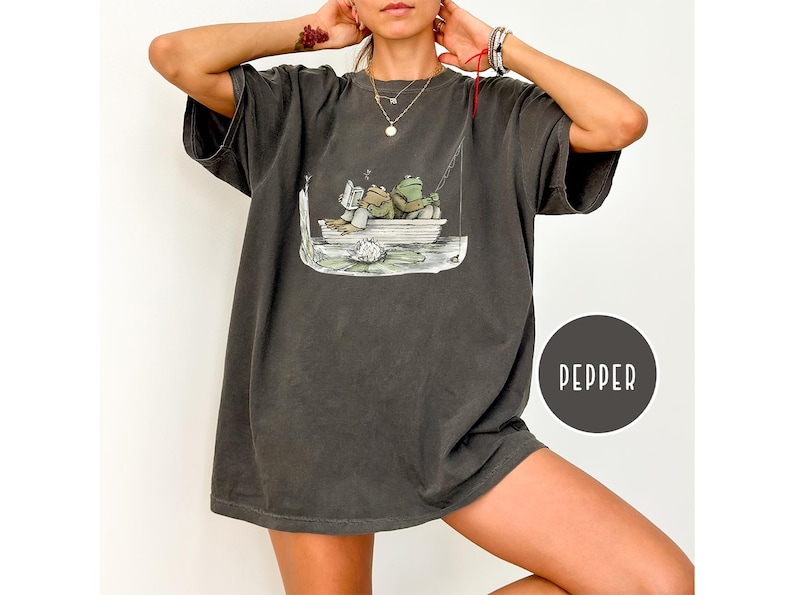Op de afbeelding: Donkergrijze oversized t-shirt met een grillige illustratie van twee kikkers in een boot, een lezend en de ander aan het vissen. De shirt heeft een relaxte pasvorm en een vintage wassing. Het woord "PEPPER" is gedrukt op een donkergrijze cirkel.