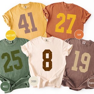 Op de afbeelding: Vijf shirts met korte mouwen in de kleuren mosterd, yam, mos, ivoor en espresso. Elk shirt heeft een groot, versleten nummer in een contrasterende kleur. De shirts zijn gerangschikt op een witte achtergrond.