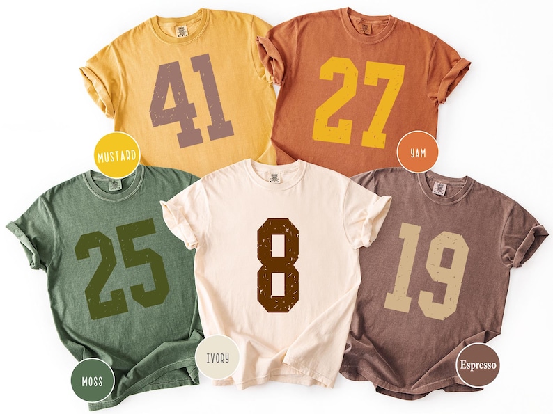 Op de afbeelding: Vijf shirts met korte mouwen in de kleuren mosterd, yam, mos, ivoor en espresso. Elk shirt heeft een groot, versleten nummer in een contrasterende kleur. De shirts zijn gerangschikt op een witte achtergrond.