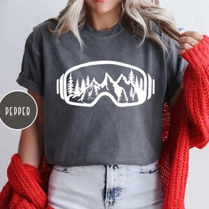 May include: Dark grey t-shirt with a white graphic of ski goggles containing a mountain and pine tree design. The word "PEPPER" is printed on a circular label on the left side of the shirt.