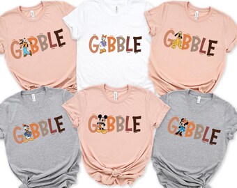 May include: A collection of six t-shirts in various colors, including pink, white, and gray. Each shirt features the word "GOBBLE" in brown and orange letters, with a different cartoon character illustration. The characters include Goofy, Daisy Duck, Pluto, Mickey Mouse, and Minnie Mouse.