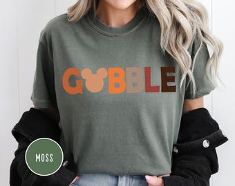 May include: Olive green t-shirt with the word "GOBBLE" in a colorful, retro font. The letter "O" is replaced with a Mickey Mouse head. The shirt is a short-sleeved, casual style.