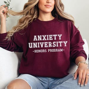 May include: A maroon sweatshirt with the text "ANXIETY UNIVERSITY - HONORS PROGRAM" in white block letters. The long-sleeved crew neck shirt is worn by a person wearing blue jeans.