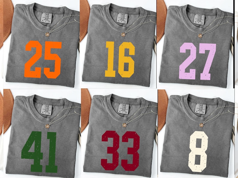Op de afbeelding: Zes grijze t-shirts met verschillende nummers in diverse kleuren: oranje, geel, lila, groen, rood en gebroken wit. Elk shirt heeft een ronde hals en een zilveren ketting.