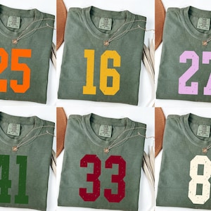 Op de afbeelding: Zes olijfgroene t-shirts met verschillende nummers in oranje, geel, lila, groen, bordeauxrood en gebroken wit. Elke shirt heeft een gouden ketting.