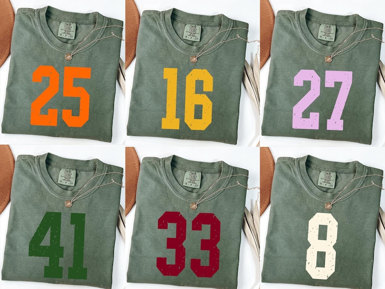 Op de afbeelding: Zes olijfgroene t-shirts met verschillende nummers in oranje, geel, lila, groen, bordeauxrood en gebroken wit. Elke shirt heeft een gouden ketting.