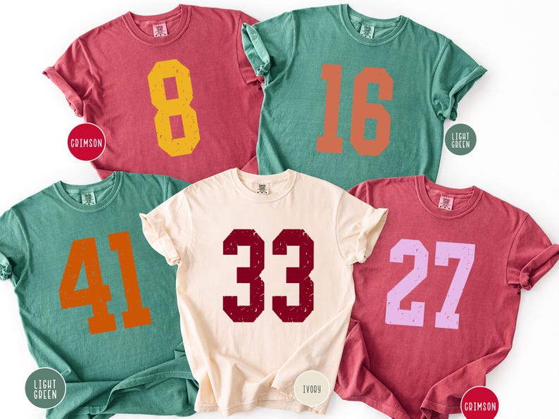 Op de afbeelding: Vijf shirts met korte mouwen in verschillende kleuren: karmozijnrood, lichtgroen en ivoor. Elk shirt heeft een groot nummer in een contrasterende kleur. Nummers zijn onder meer 8, 16, 41, 33 en 27. De shirts hebben een vintage, versleten uitstraling.
