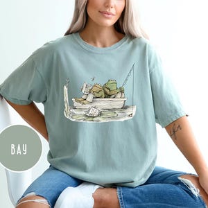 Op de afbeelding: Lichtblauw T-shirt met een illustratie van twee kikkers in een boot. E&eacute;n kikker leest, de ander vist. Het T-shirt heeft een losse pasvorm en is gemaakt van zacht materiaal.