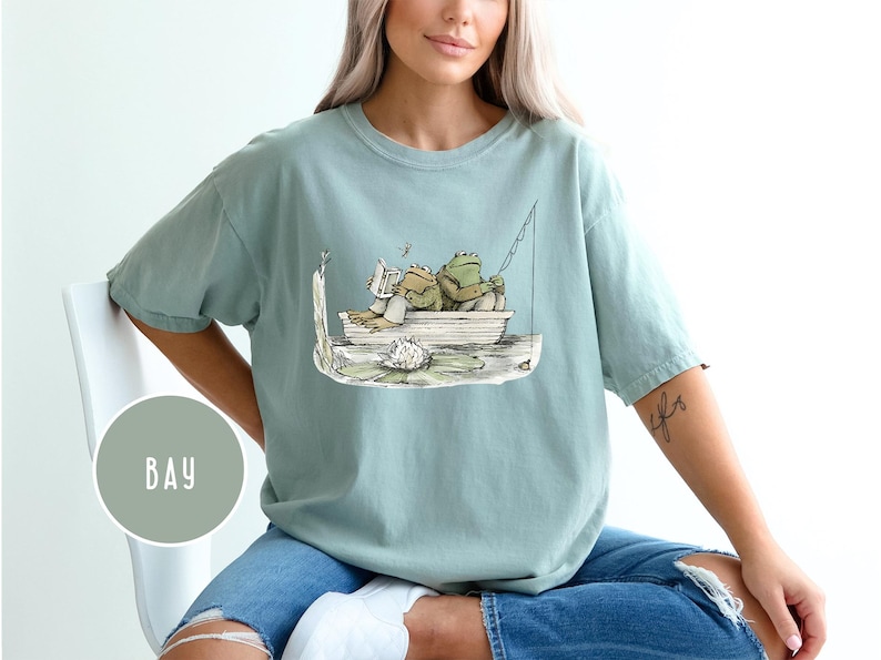 Op de afbeelding: Lichtblauw T-shirt met een illustratie van twee kikkers in een boot. E&eacute;n kikker leest, de ander vist. Het T-shirt heeft een losse pasvorm en is gemaakt van zacht materiaal.