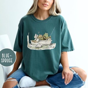 Op de afbeelding: Blauwgroen T-shirt met een cartoon illustratie van twee kikkers in een boot, een leest en de ander vist. Het ontwerp bevat een waterlelie en de tekst "BLUE-SPRUCE" in een cirkel.