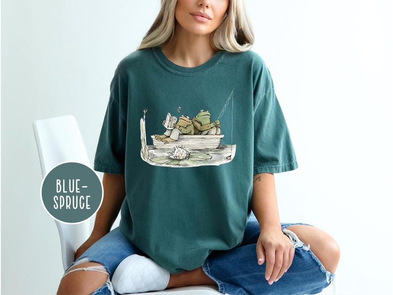 Op de afbeelding: Blauwgroen T-shirt met een cartoon illustratie van twee kikkers in een boot, een leest en de ander vist. Het ontwerp bevat een waterlelie en de tekst "BLUE-SPRUCE" in een cirkel.