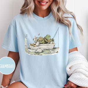 Op de afbeelding: Lichtblauw T-shirt met een grillige illustratie van twee kikkers in een boot. De ene kikker leest een boek, de andere vist. Een libelle zweeft in de buurt. Het ontwerp bevat een waterlelie en een hengel.