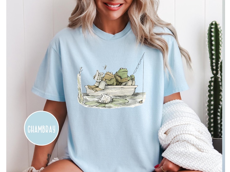 Op de afbeelding: Lichtblauw T-shirt met een grillige illustratie van twee kikkers in een boot. De ene kikker leest een boek, de andere vist. Een libelle zweeft in de buurt. Het ontwerp bevat een waterlelie en een hengel.