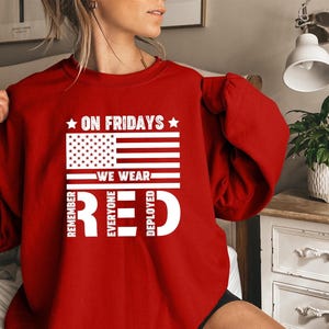 May include: Red sweatshirt with a white graphic of the American flag and the text "ON FRIDAYS WE WEAR RED". The text also includes "REMEMBER EVERYONE DEPLOYED". The sweatshirt is a solid red color.