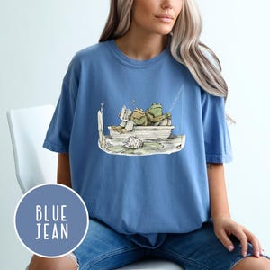 Op de afbeelding: Blauw spijkerbroek T-shirt met een grillige illustratie van twee kikkers in een boot, de ene leest en de andere vist. Het ontwerp bevat een waterlelie en een vislijn. Het shirt heeft een ronde hals en korte mouwen.