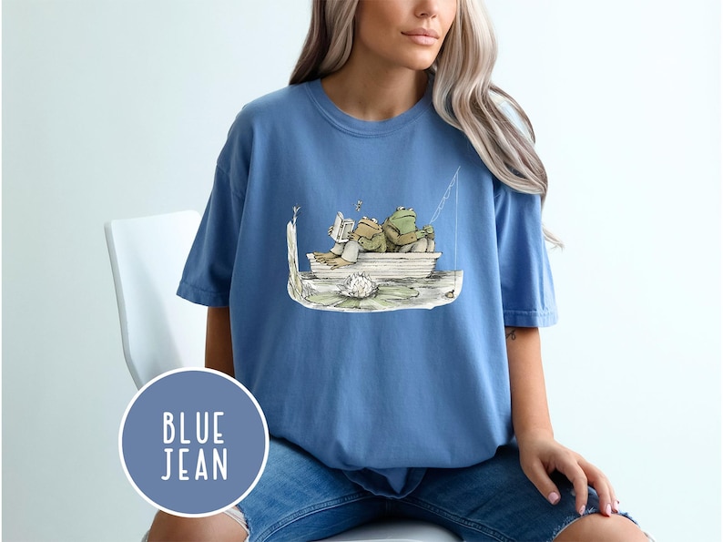 Op de afbeelding: Blauw spijkerbroek T-shirt met een grillige illustratie van twee kikkers in een boot, de ene leest en de andere vist. Het ontwerp bevat een waterlelie en een vislijn. Het shirt heeft een ronde hals en korte mouwen.