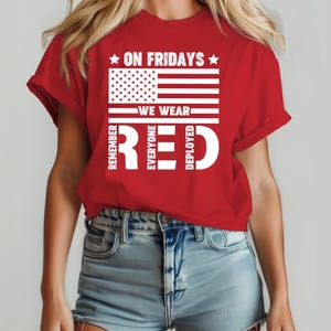 May include: A red t-shirt with a white graphic of the American flag and the text "ON FRIDAYS WE WEAR RED REMEMBER EVERYONE DEPLOYED". The shirt has short sleeves and a crew neck.