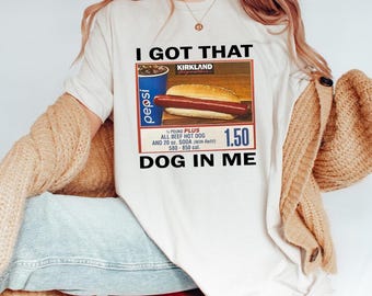 May include: Cream-colored t-shirt with the text "I GOT THAT DOG IN ME." The shirt features a graphic of a hot dog, soda, and the Kirkland logo. The graphic includes the text "¼ POUND PLUS ALL BEEF HOT DOG AND 20 oz. SODA (with Refill) 580-850 cal."