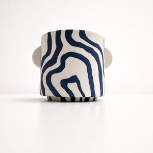 Könnte beinhalten: Ein Keramik-Pflanztopf mit cremefarbenem Sockel und abstraktem, marineblauem Wellenmuster. Der Pflanztopf hat zwei kleine, abgerundete Griffe und einen leicht erhöhten Sockel. Ein modernes Dekorationsobjekt.