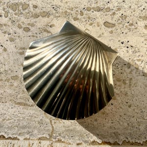 Może przedstawiać: Srebrny, dekoracyjny przedmiot w kształcie muszli. Muszla ma prążkowaną fakturę i zakrzywiony kształt, z gładką, odblaskową powierzchnią. Obiekt jest prawdopodobnie wykonany z metalu i jest prezentowany na teksturowanej, jasnej powierzchni.