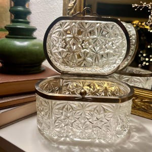 Peut inclure: Une boîte à bijoux en verre transparent d'époque, ornée d'un motif en étoile. La boîte ovale est dotée d'un couvercle à charnière et d'un cadre en métal. La boîte est ouverte, révélant l'intérieur. Une lampe verte et un miroir doré sont en arrière-plan.
