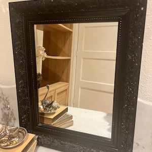 Könnte beinhalten: Quadratischer Spiegel mit schwarzem Rahmen und aufwendigen Details. Der Spiegel spiegelt einen Raum mit einem Holzschrank und einer weißen Tür wider. Dekorative Gegenstände sind in der Reflexion und vor dem Spiegel sichtbar.