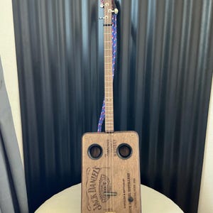 Może przedstawiać: Ręcznie wykonana gitara z pudełka po cygarach z drewnianym korpusem z logo i tekstem Jack Daniel's. Instrument ma długą szyjkę, struny i niebiesko-czerwony pasek. Tło to czarna blacha falista.