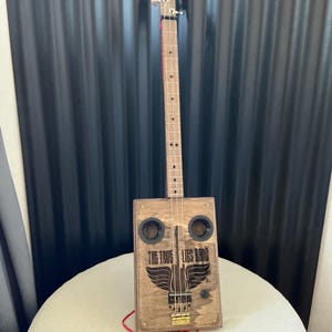 Könnte beinhalten: Eine handgefertigte Zigarrenkistengitarre mit einem Korpus und Hals aus Holz. Auf der Vorderseite der Kiste steht der Text "THE TRUE LIES BAND" über einer Grafik eines Vogels mit ausgebreiteten Flügeln. Die Gitarre hat vier Saiten und zwei kreisförmige Schalllöcher.