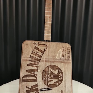 Könnte beinhalten: Eine handgefertigte Zigarrenkistengitarre mit einem Holzhals und einem Korpus aus einer Jack Daniel's Whiskey-Box. Die Gitarre zeigt das Jack Daniel's Logo und Text, mit einer roten Schnur und einem kleinen Anhänger.