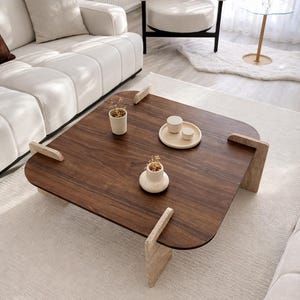 May include: A modern living room scene featuring a dark wood coffee table with light-colored stone legs. The table is adorned with small decorative items. A white sofa and armchair are in the background, set on a light-colored rug.