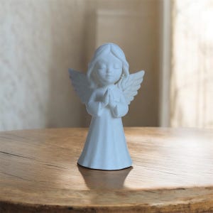 Op de afbeelding: Een wit engelenbeeldje met gevouwen handen en gesloten ogen. De engel heeft gedetailleerde vleugels en golvend haar. Het beeldje staat op een houten oppervlak.
