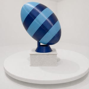 Könnte beinhalten: Eine blau und hellblau gestreifte, rugbyballförmige Skulptur. Die Skulptur ist auf einem dunkelblauen Ständer auf einem kleinen, weißen ziegelartigen Sockel montiert. Das Objekt befindet sich vor einem weißen Hintergrund.