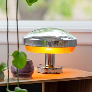 May include: A chrome and orange mushroom-shaped table lamp. The lamp has a reflective, domed top and a cylindrical base. The orange section of the lamp glows. The lamp sits on a wooden surface, with a small potted plant and a purple ceramic pot in the background.