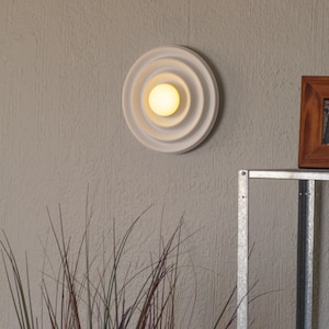May include: A white, circular wall sconce with concentric rings and a central, glowing light. The fixture is mounted on a textured, light gray wall. A wooden framed picture and metal shelf are visible.
