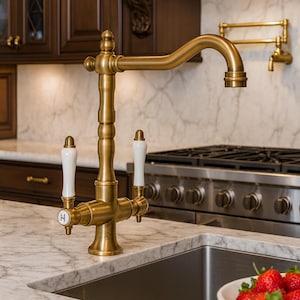 Könnte beinhalten: Eine goldfarbene Küchenarmatur mit weißen Griffen und eine passende Wandarmatur. Der Wasserhahn ist auf einer Marmorarbeitsplatte mit einem Edelstahlbecken installiert. Dunkle Holzschränke und ein Gasherd sind im Hintergrund zu sehen.