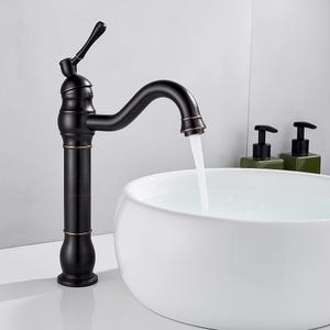 May include: A tall, matte black bathroom tap with a curved spout and a single lever handle, featuring subtle copper-toned accents at the joints. Water flows from the spout into a white, round, vessel-style sink basin, creating a clear stream.