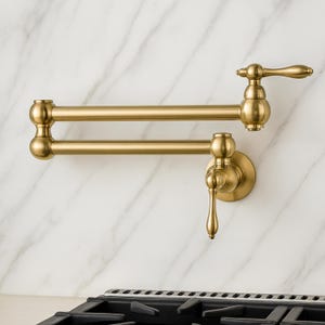May include: A gold-colored, wall-mounted pot filler faucet with a double-jointed arm. The faucet has a classic design with a lever handle and a round base. The fixture is mounted on a white marble-patterned wall, above a black stovetop.