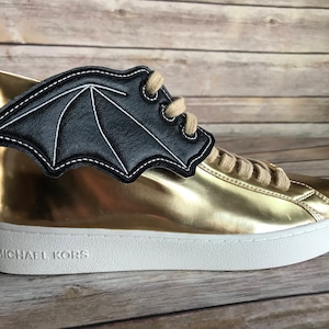 May include: Gold high-top sneakers with a black bat wing detail and a white sole. The sneakers have a Michael Kors logo on the side.