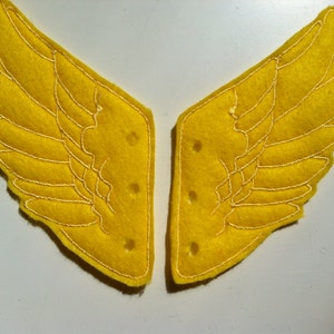 May include: A pair of yellow felt wings with white stitching, each wing has a rounded shape and a design of feathers. The wings are sewn together at the top.