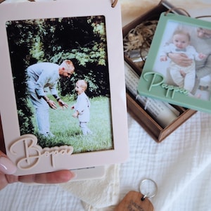 May include: Two photo frames, one light pink and one light green, each with a photo inside. The pink frame has the word "Bappa" in cursive, and the green frame has the word "Pappa". Both frames have a leather cord for hanging.