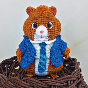 Op de afbeelding: Een handgemaakte gehaakte hamsterpop met een blauwe blazer, wit overhemd en gestreepte stropdas. De hamster heeft bruine vacht, een wit gezicht en grote ogen. Hij zit in een gevlochten mand. Een uniek, handgemaakt item.