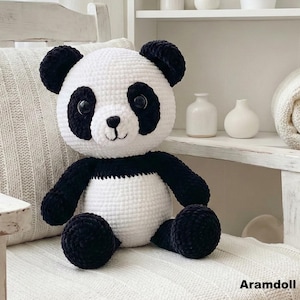 Puede incluir: Un oso panda de ganchillo hecho a mano, en blanco y negro. El panda tiene orejas, ojos, brazos y piernas negros, con un cuerpo blanco. El panda está sentado en una silla blanca. El texto "Aramdoll" está en la esquina inferior derecha.