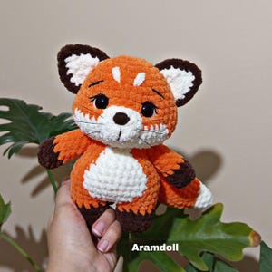 Puede incluir: Un peluche de zorro de ganchillo naranja y blanco hecho a mano con detalles marrones. El zorro tiene grandes ojos negros, un pecho blanco y una cola esponjosa. El peluche se sostiene frente a una hoja verde.