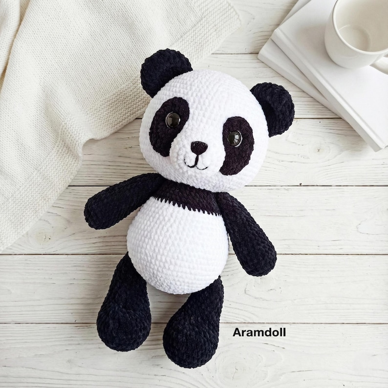 May include: A handmade panda bear plush toy with black and white coloring. The panda has black ears, eye patches, arms, and legs, with a white body. The plush is sitting on a light wood surface. The text "Aramdoll" is visible.
