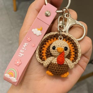 May include: A handmade turkey keychain with a pink strap. The turkey is crocheted with brown, orange, and beige yarn, featuring a red wattle and black eyes. The strap has the word "wish" and decorative elements.