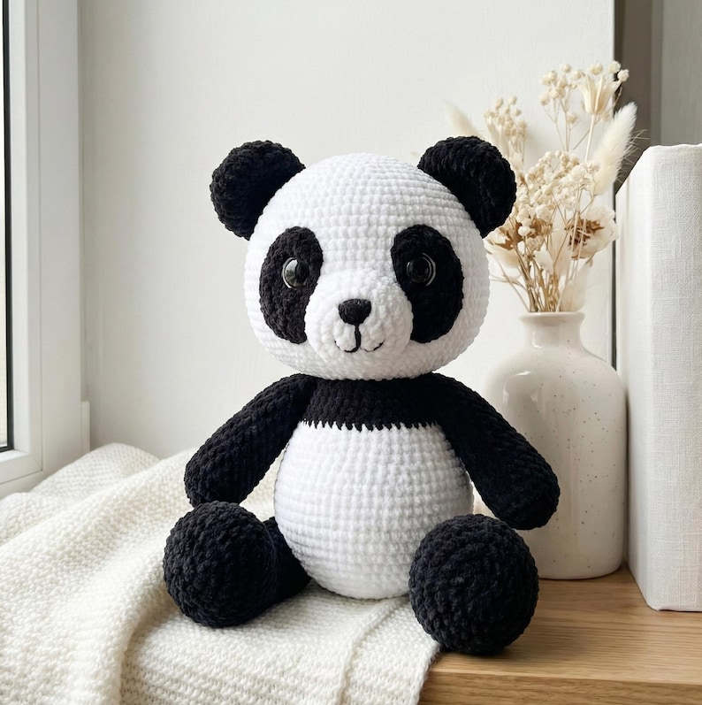 May include: A handmade crochet panda bear with black and white yarn. The panda has black ears, eyes, arms, and legs, with a white head and body. The stuffed animal is sitting on a white blanket.
