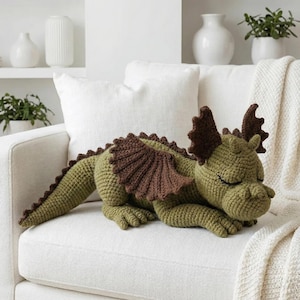 May include: A handmade, crochet dragon in shades of green and brown, resting on a white sofa. The dragon features detailed wings, a spiky spine, and closed eyes, suggesting a sleeping pose. The soft toy is a decorative item.